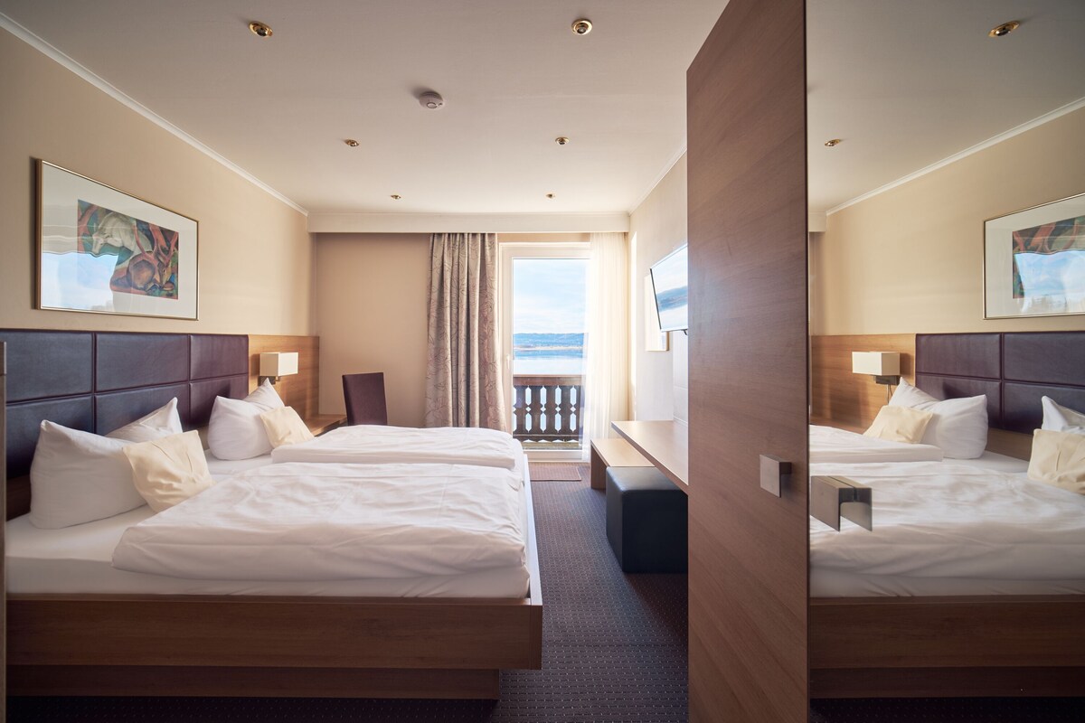 A hotel room with two beds, a desk, a view of the sea, and artwork on the walls.