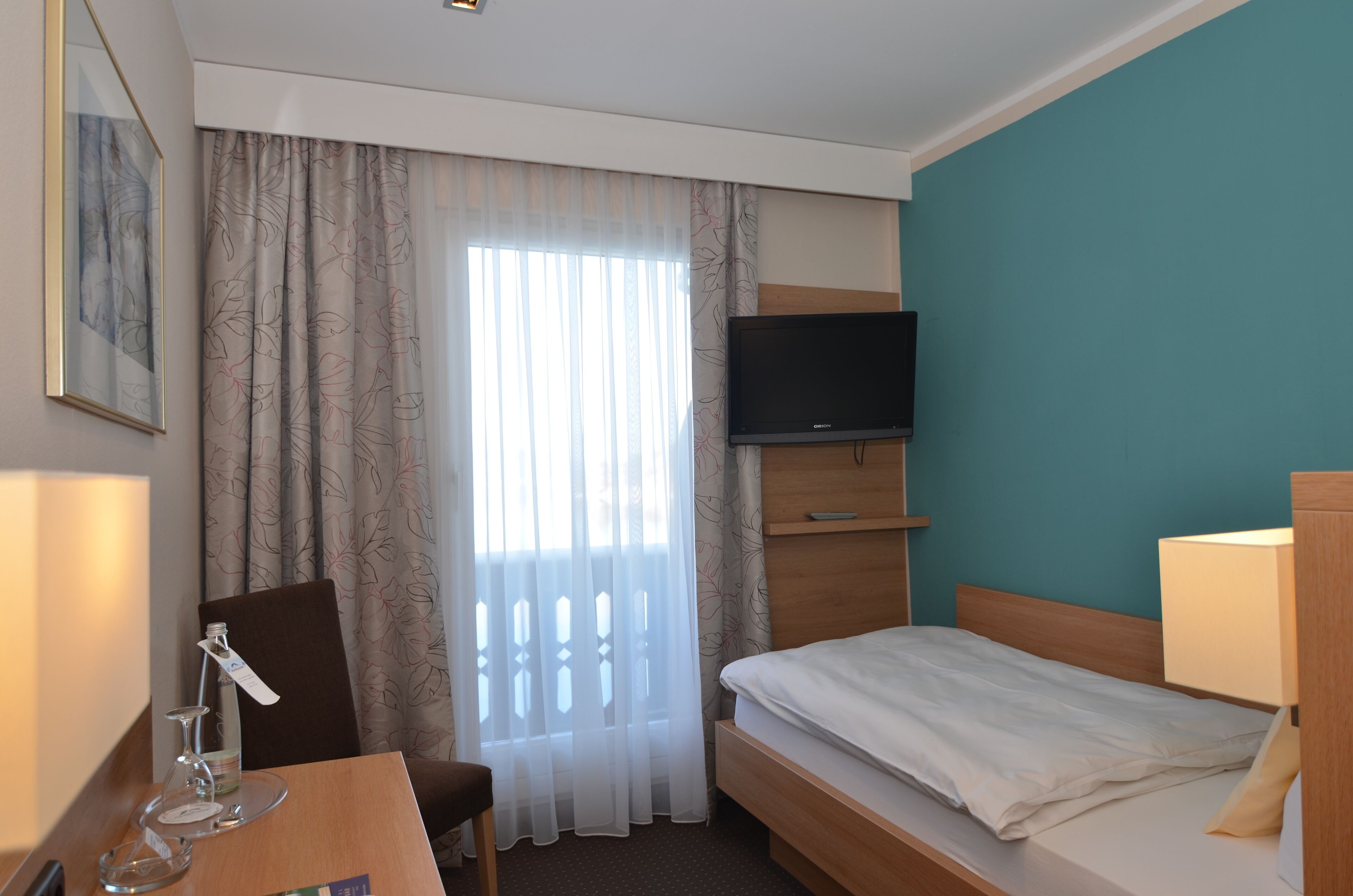Een hotelkamer met een bed, een bureau met een stoel, een aan de muur bevestigde televisie en een raam met dunne gordijnen.