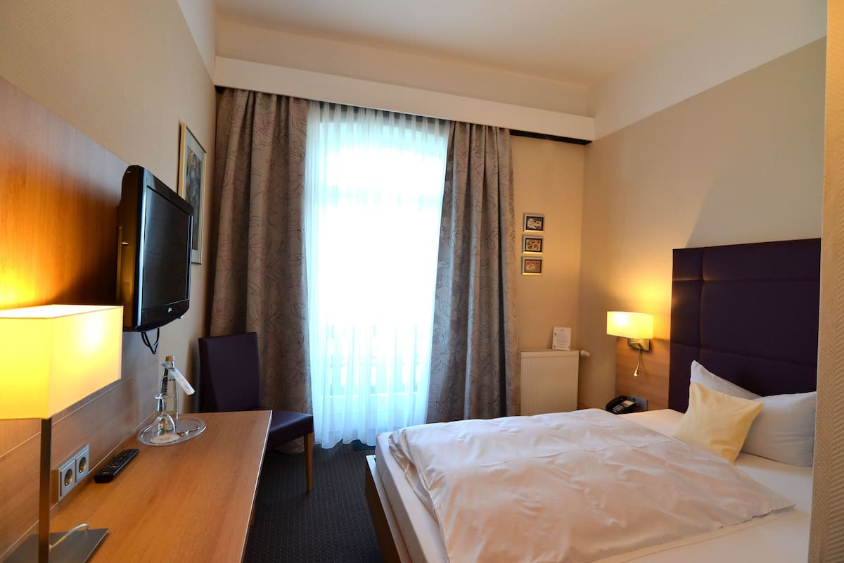 Quarto de hotel com cama, mesa de cabeceira, luminária, televisão e cortinas.