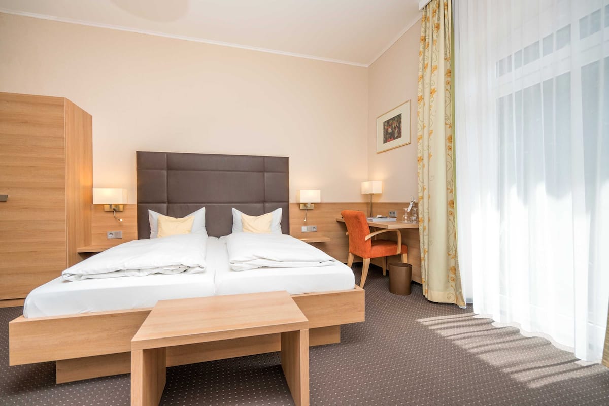 Quarto de hotel com cama de casal, mesas de cabeceira com luminárias, escrivaninha com cadeira e janela com cortinas.