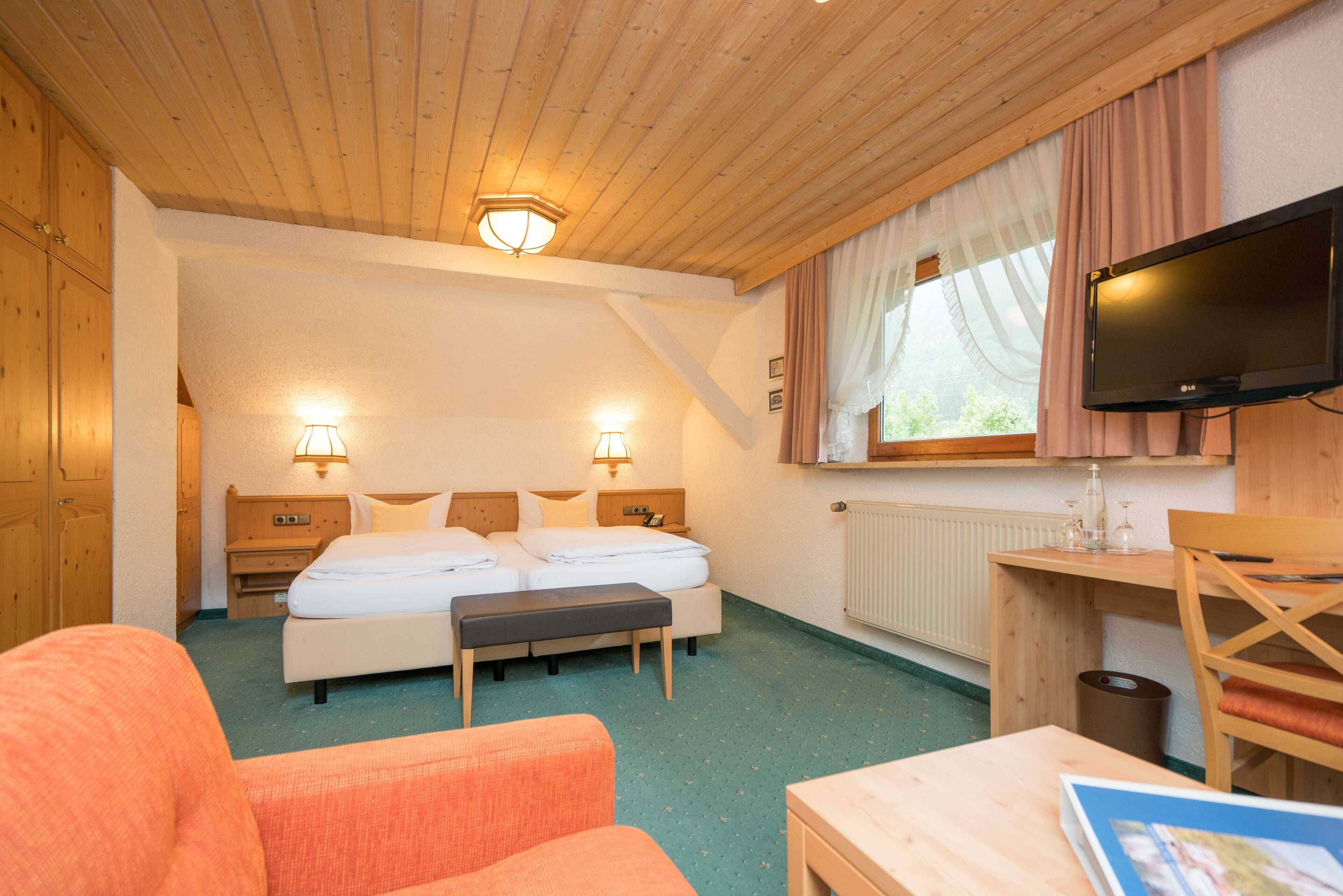 A hotel room with a wooden ceiling, a bed with two pillows, a desk with a television, and a window with curtains.