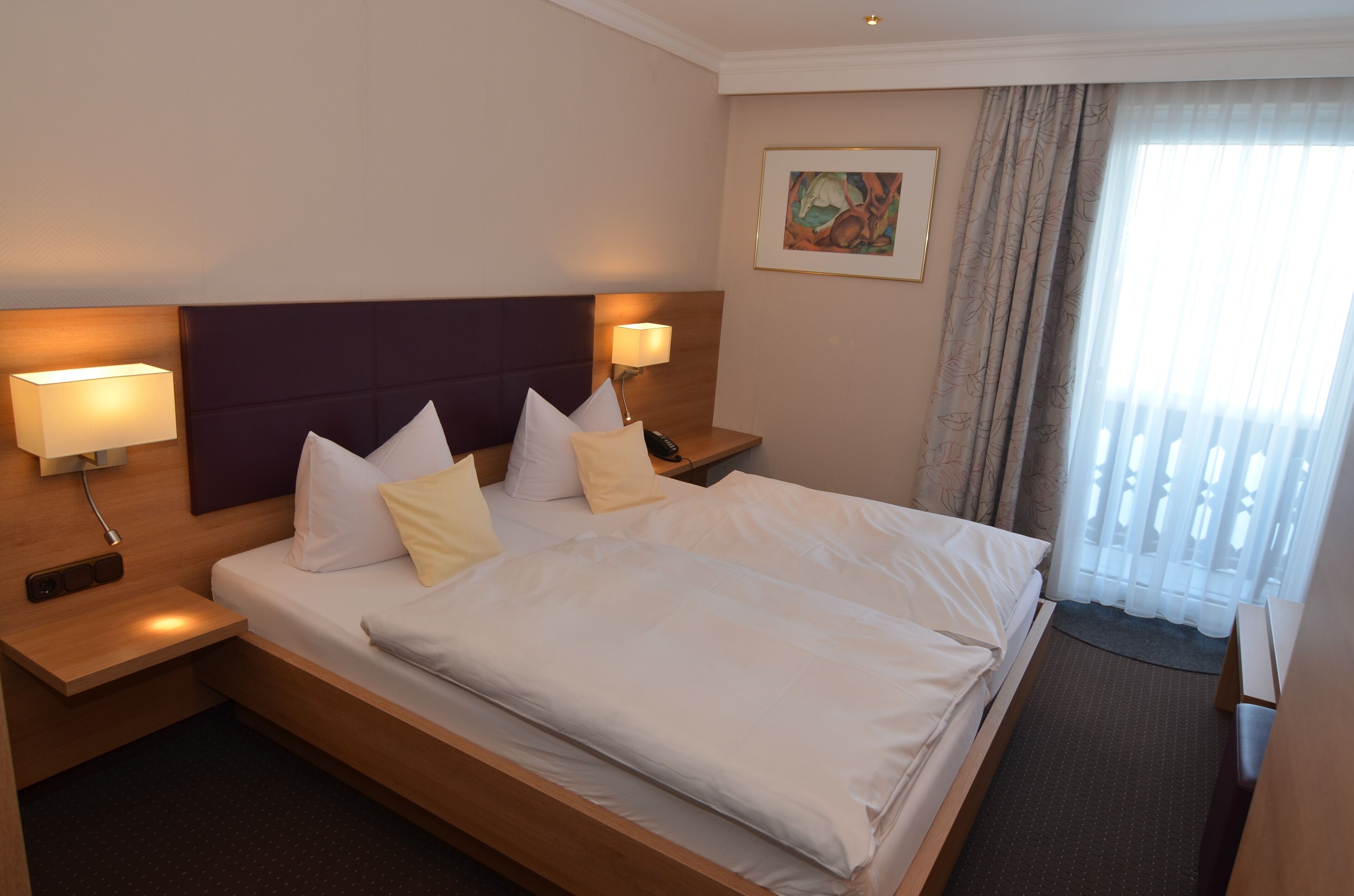 Habitación de hotel con una cama grande, dos mesitas de noche con lámparas, un cabecero, un cuadro en la pared y una ventana con cortinas.