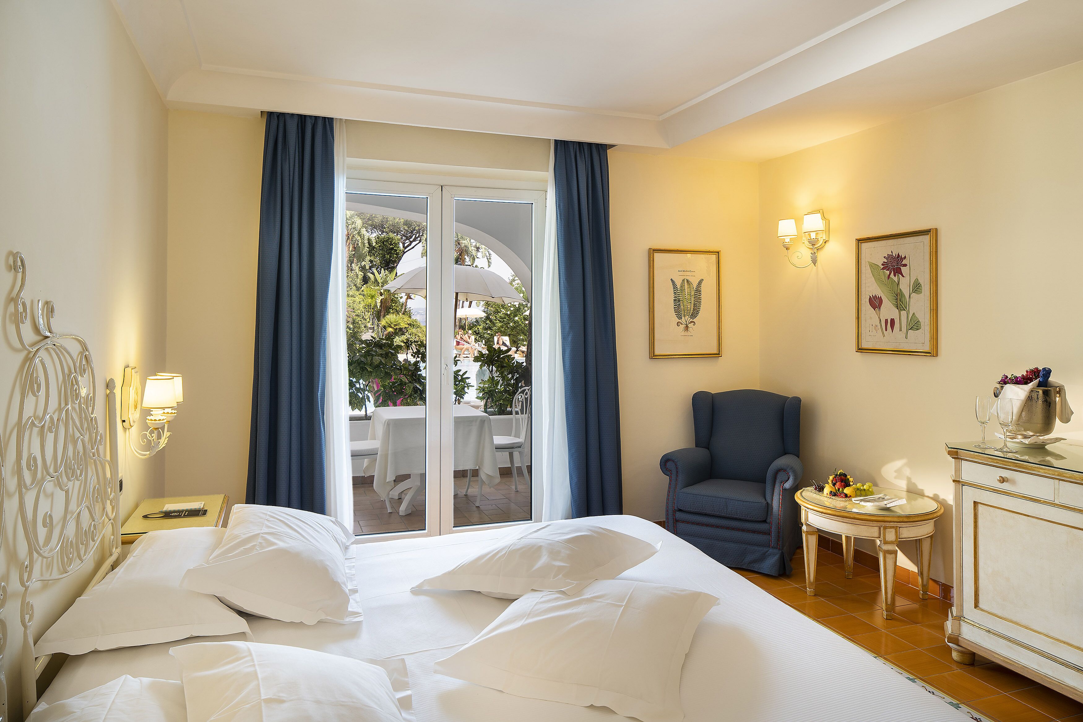 A hotel room with a bed, a chair, a small table with a vase, and a view of the outdoors.