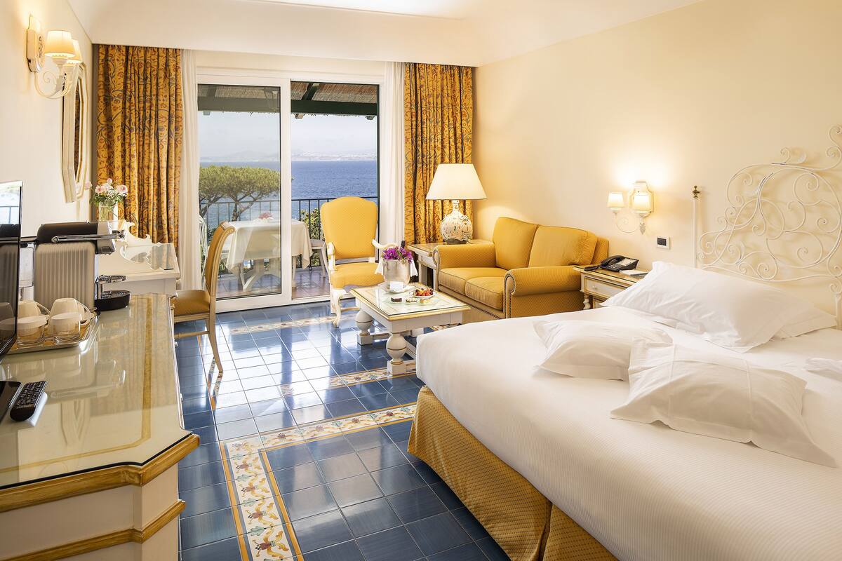 Ein Hotelzimmer mit einem Bett, einem Schreibtisch mit Computer, einem Sessel, einem kleinen Tisch mit Lampe und Blick aufs Meer.