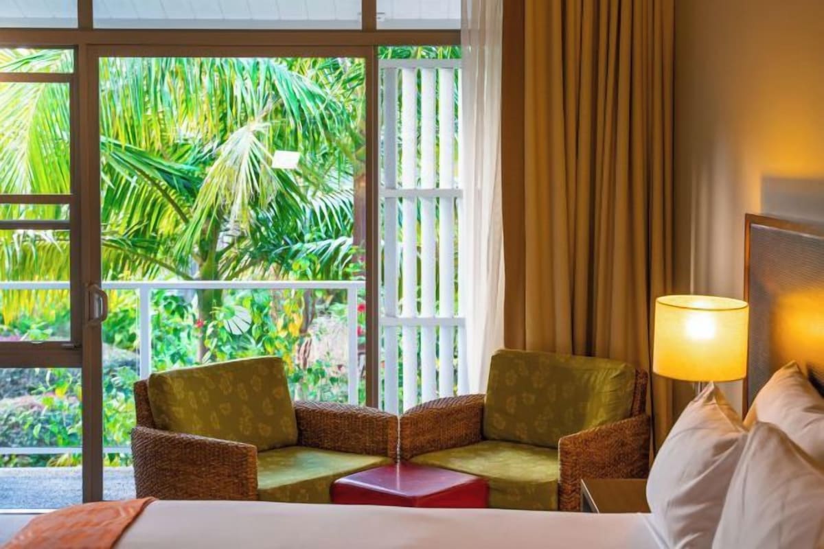 A hotel room with a bed, two chairs, a small table, a lamp, and a view of lush greenery through a window.