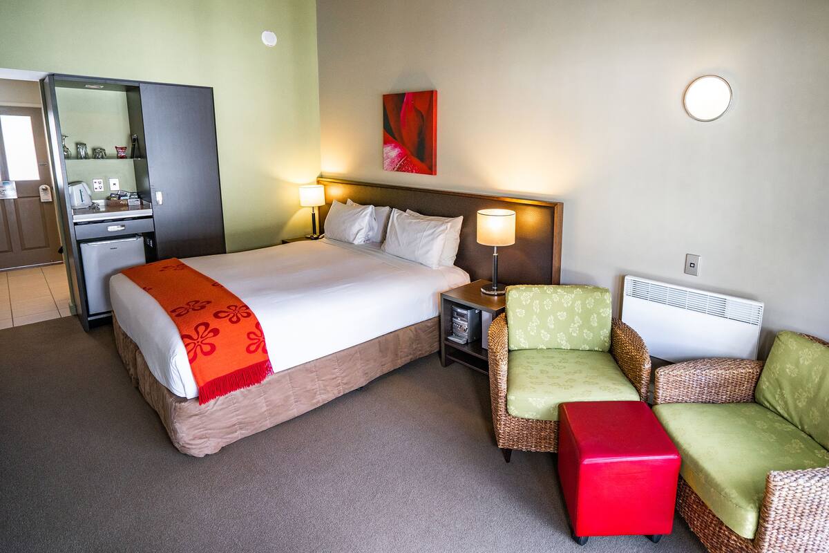 A hotel room with a bed, a desk, a chair, a red ottoman, and a wall-mounted heater.
