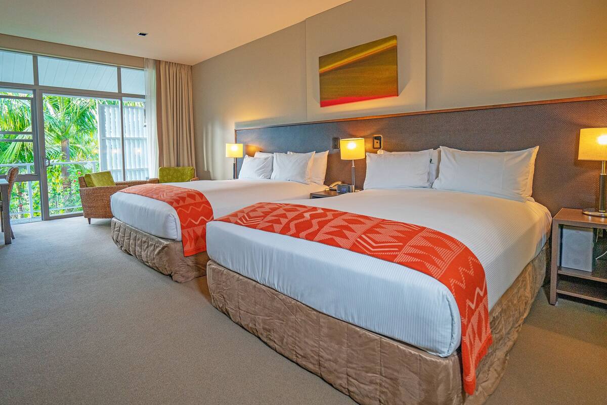 A hotel room with two beds, a sitting area with a chair, and a view of greenery through the window.