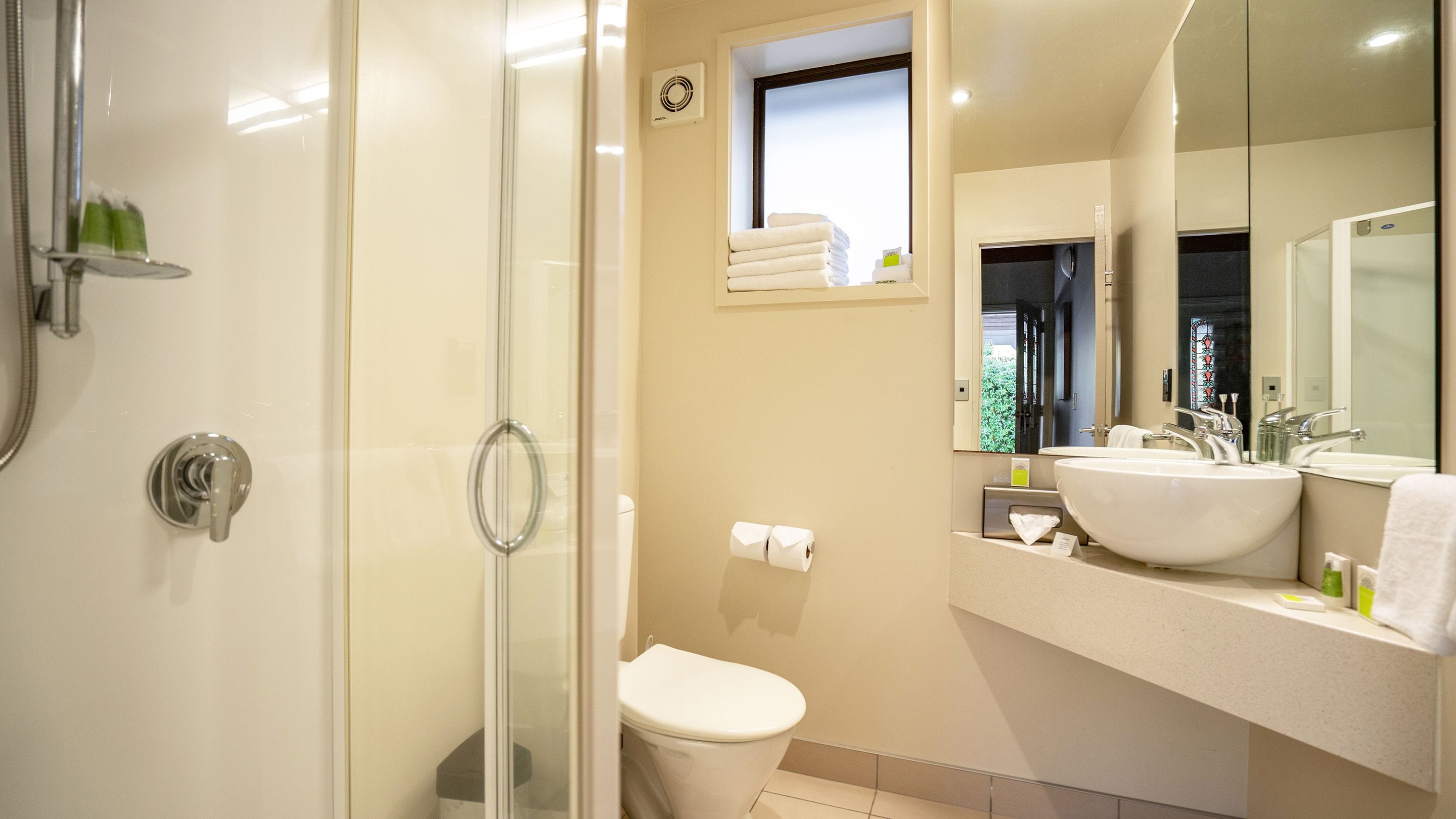 A modern bathroom with a glass shower enclosure, a toilet, a sink with a mirror, and a window providing natural light.