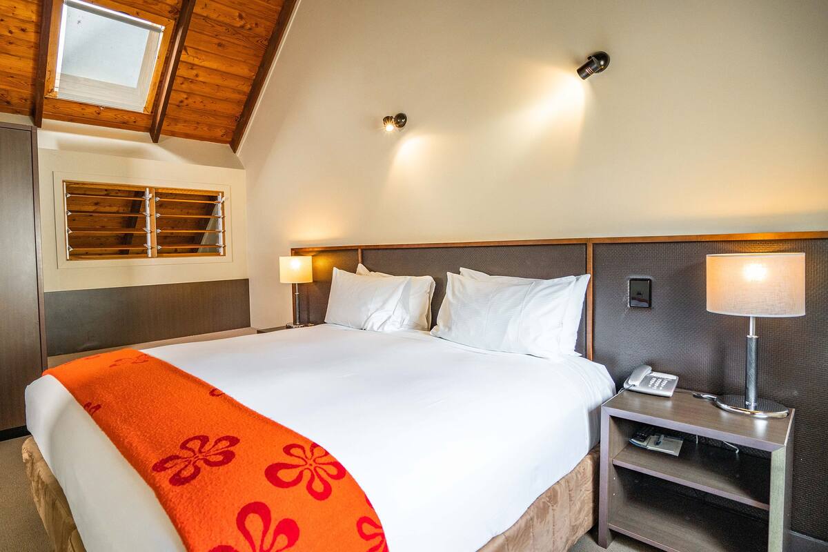 A hotel room with a large bed, a nightstand with a lamp, a wardrobe, and a window with blinds.