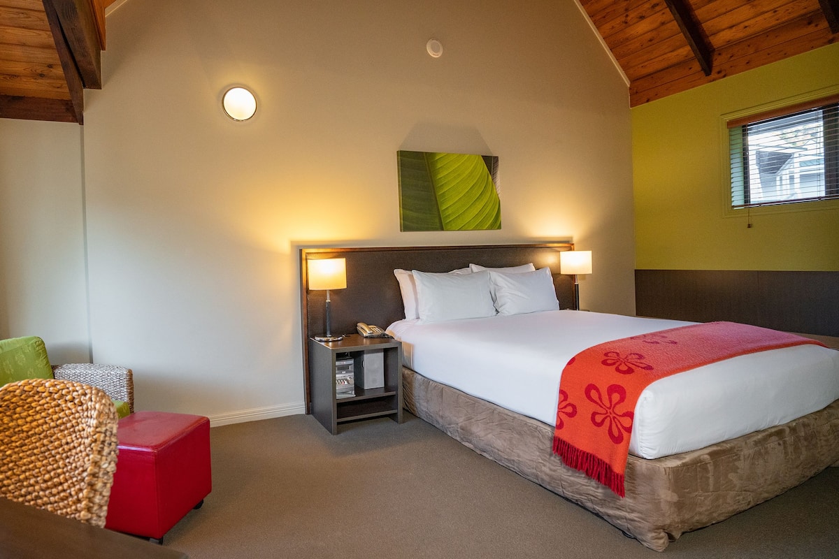 A hotel room with a large bed, a nightstand with a lamp, a red ottoman, a green chair, and a window with blinds.