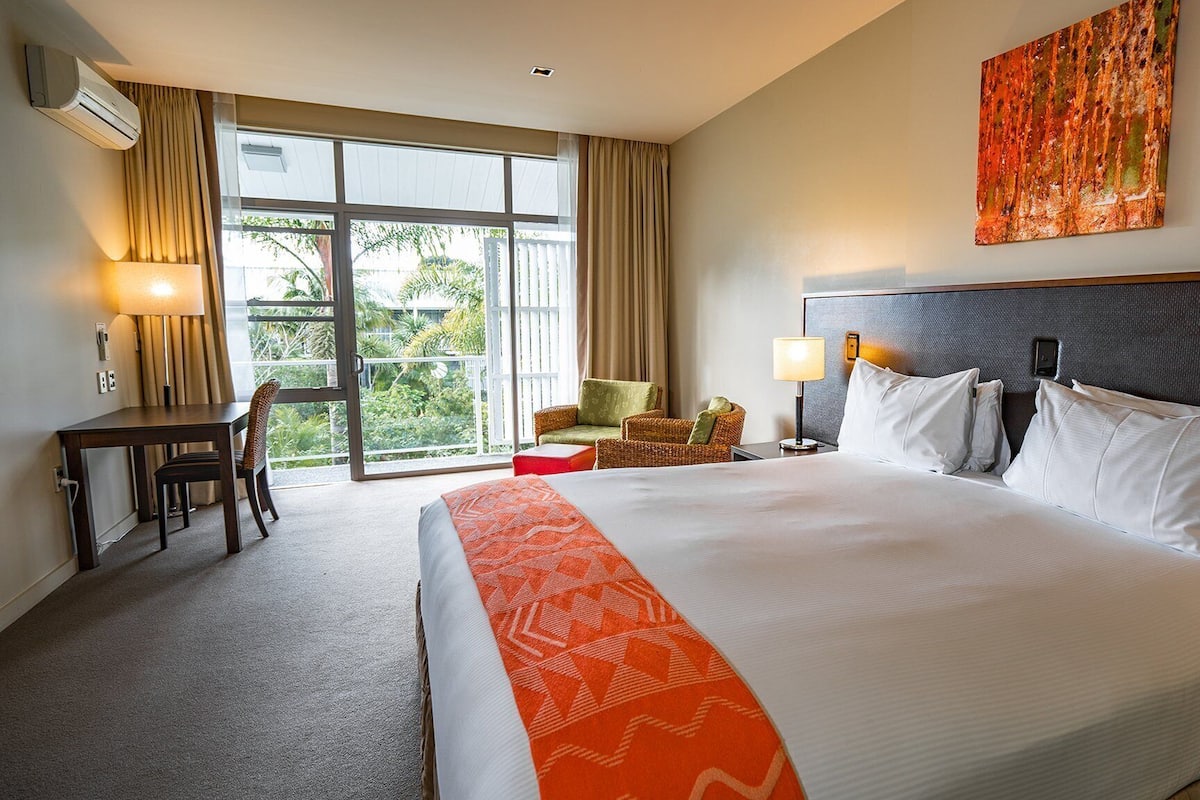 A hotel room with a large bed, a desk, a chair, and a view of greenery through the window.