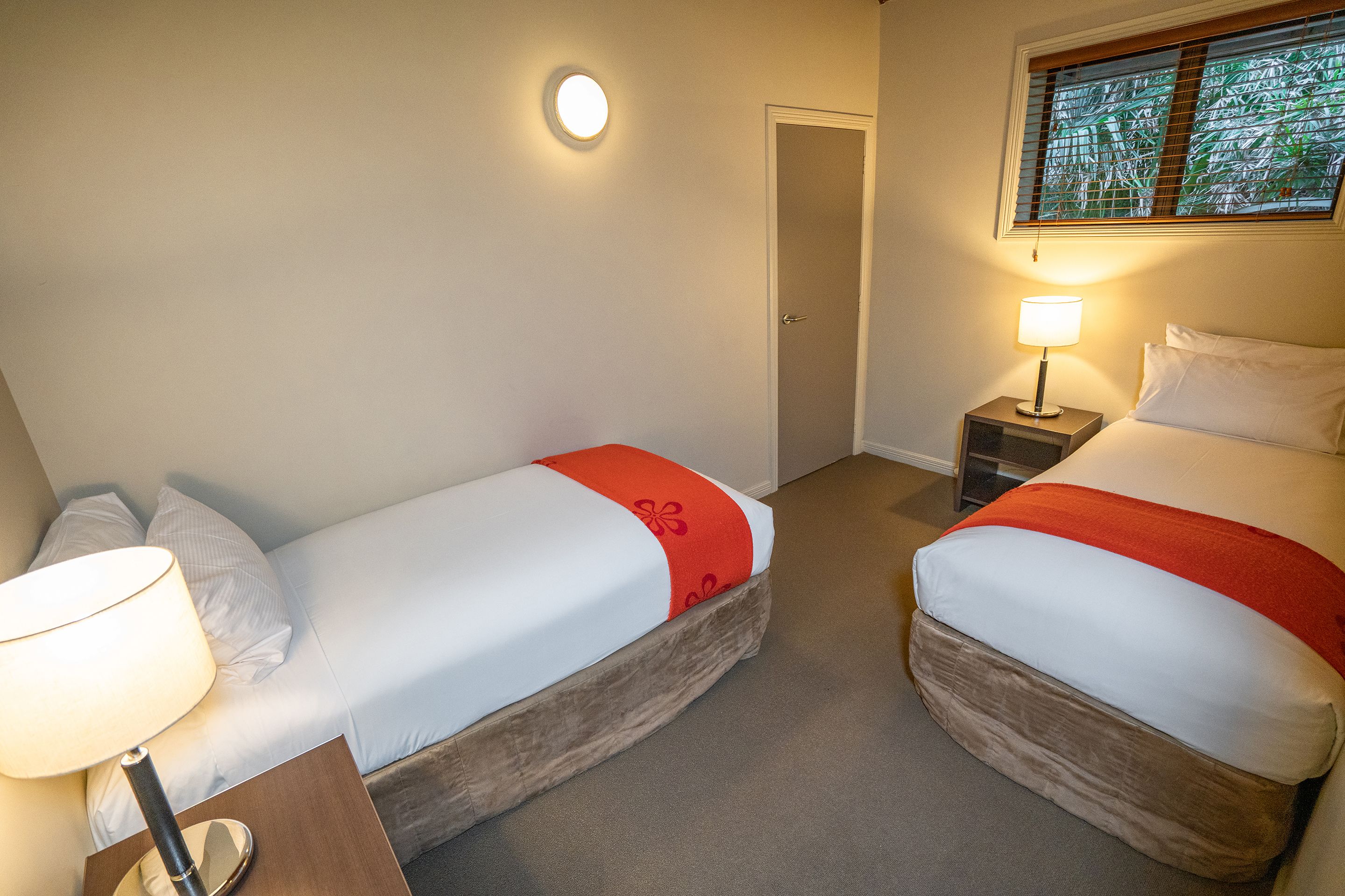 A hotel room with two beds, a nightstand with a lamp, a door, and a window with blinds.