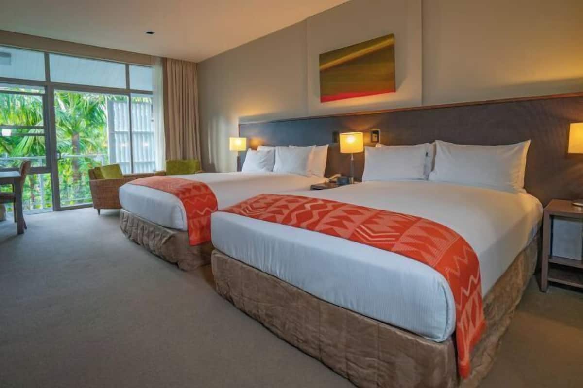 A hotel room with two beds, a desk, and a view of greenery outside.