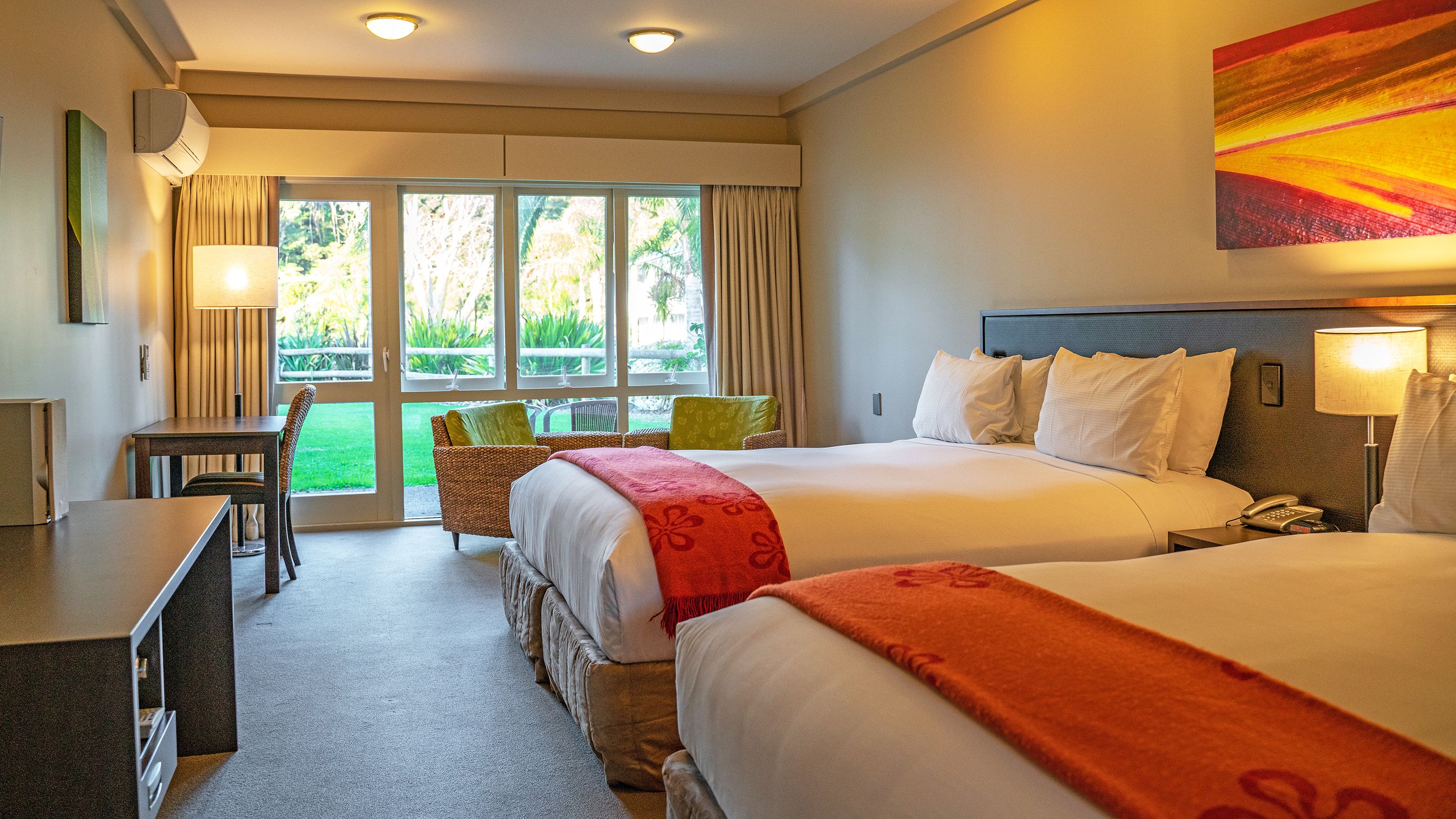 Ein Hotelzimmer mit zwei Betten, einem Schreibtisch und einem Stuhl. Durch die Fenster ist Grün anzusehen.