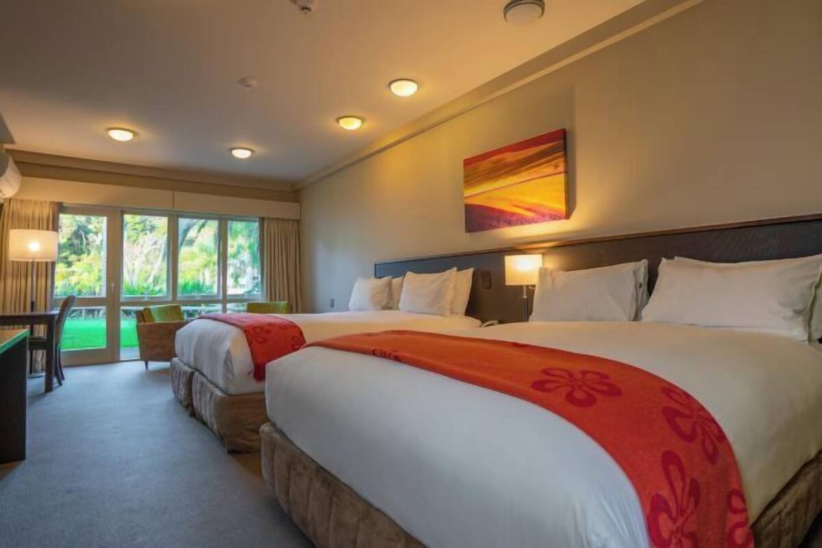 A hotel room with two beds, a desk, and a view of greenery through the windows.
