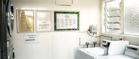 A laundry area with a washing machine, a dryer, and a window with frosted glass. There are signs on the wall providing information about the laundry process and costs.