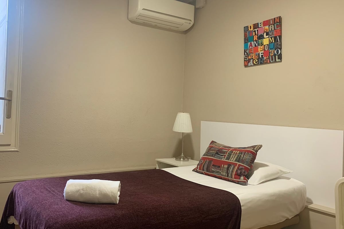 A hotel room with a bed, a nightstand, a lamp, and a window with a view of buildings outside.