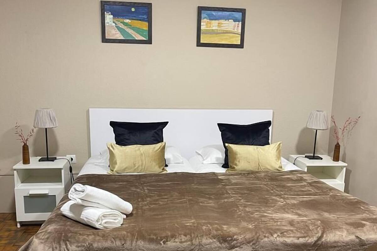 A neatly made bed with a brown bedspread, two pillows, and a folded towel on a wooden floor. There are two bedside tables with lamps and a nightstand with a lamp. Two framed pictures hang on the wall above the bed.