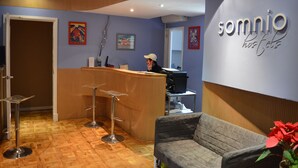 A reception area with a wooden reception desk, two bar stools, a grey sofa, and a wall with the name 'somnio hostels'. - Somnio Hostels (Barcelona)