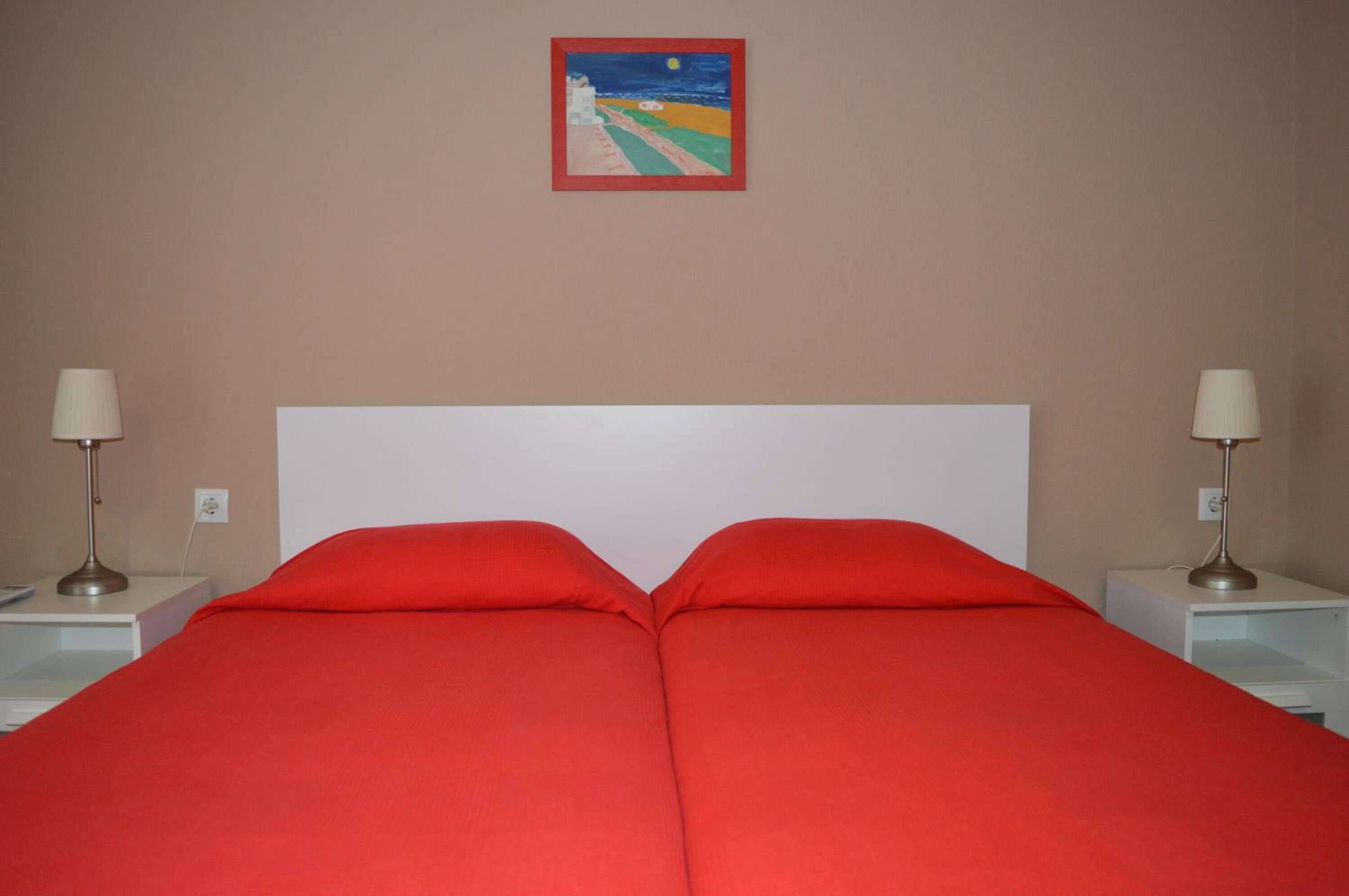 Una cama con ropa de cama roja, cabecera blanca y dos mesitas de noche con lámparas.