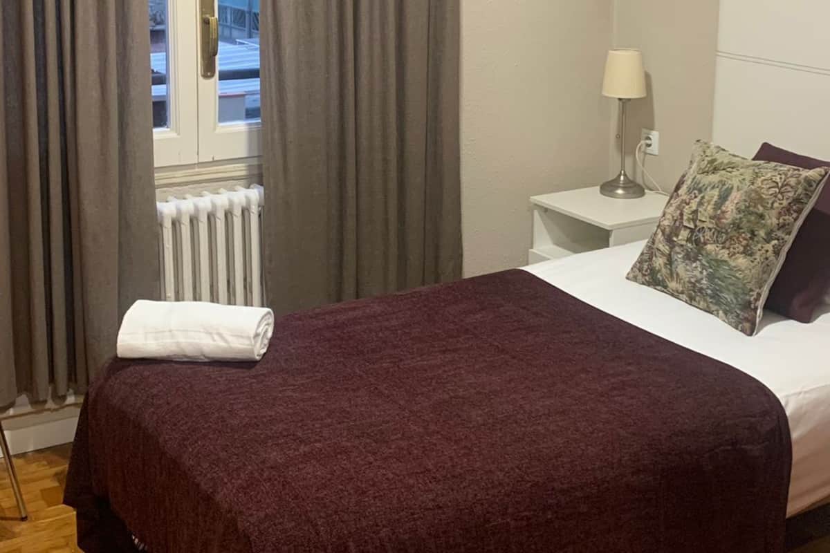 A bedroom with a bed, a nightstand, a lamp, and a window with curtains.