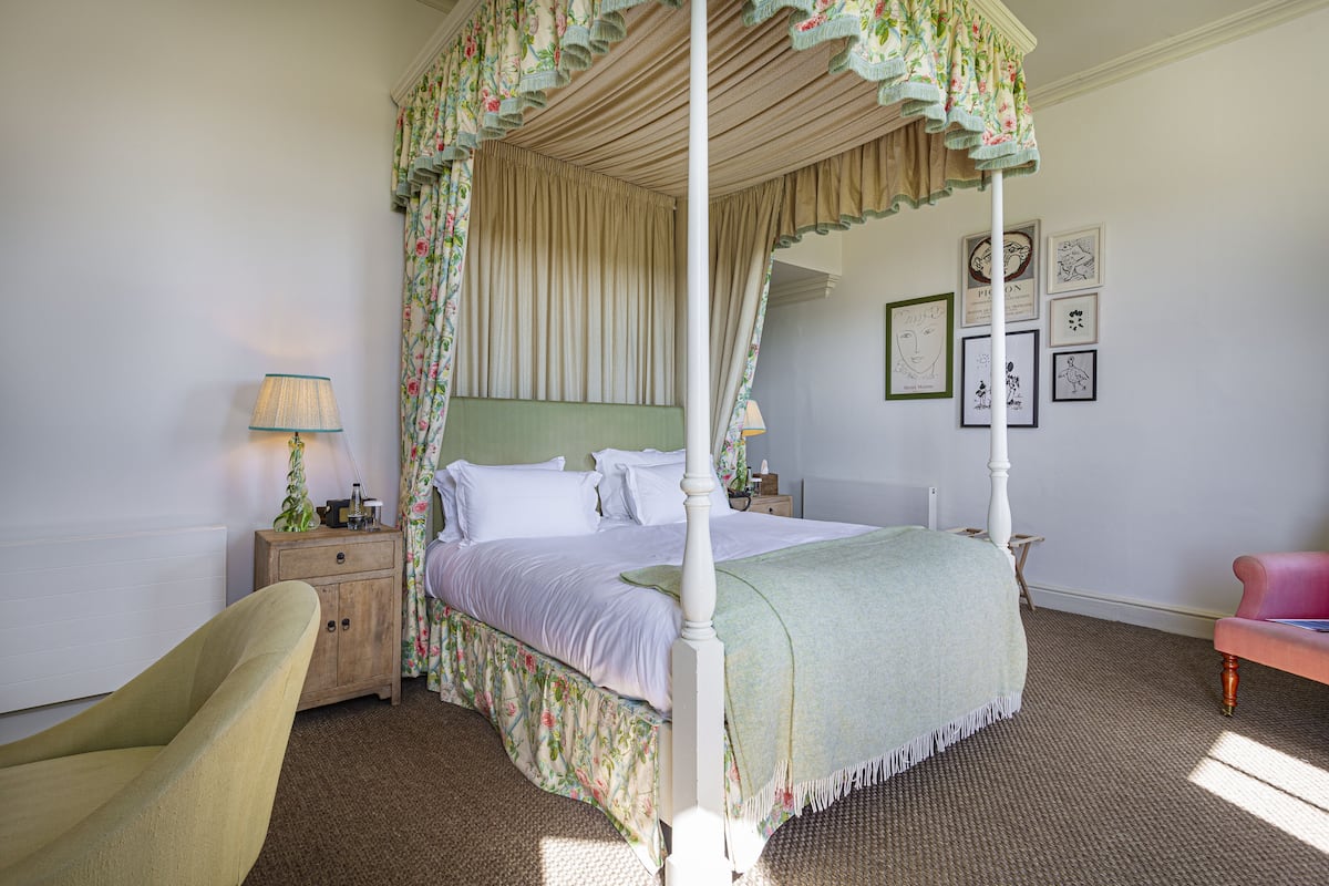 Ein Himmelbett mit Vorhängen mit Blumenmuster, ein Nachttisch mit Lampe und ein Sessel.