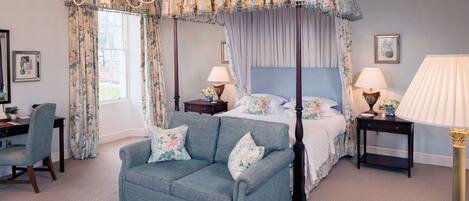 A four-poster canopy bed with floral curtains, a blue sofa, a desk with a chair, and two bedside tables with lamps.