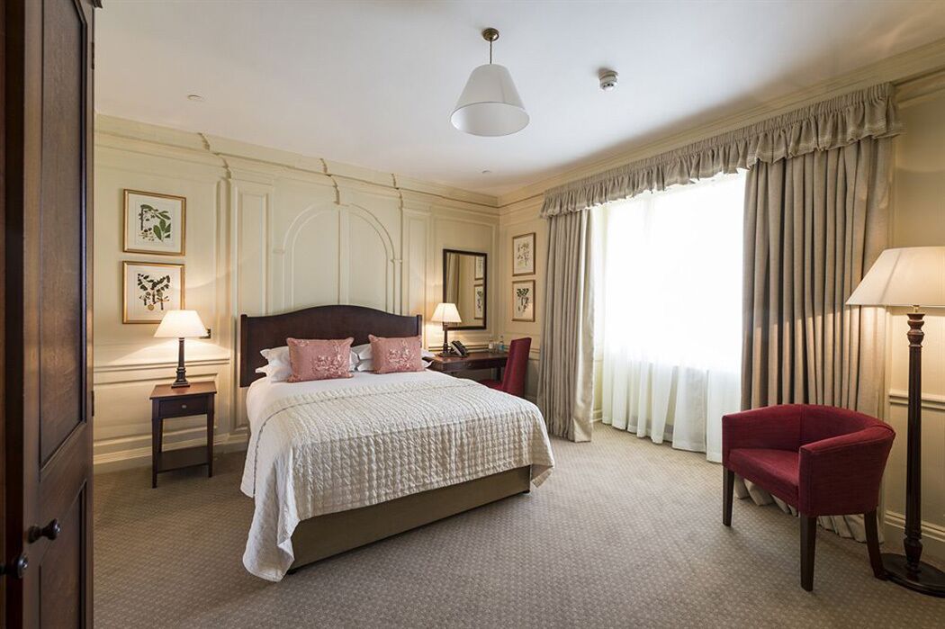 A hotel room with a large bed, two bedside tables, a red armchair, a desk with a chair, and a large window with curtains.