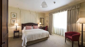 Une chambre d’hôtel avec un grand lit, deux tables de chevet, un fauteuil rouge, un bureau avec une chaise et une grande fenêtre avec des rideaux.