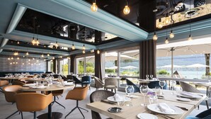 Un restaurante con varias mesas dispuestas para comer, amplios ventanales que ofrecen vistas al exterior y lámparas colgantes.