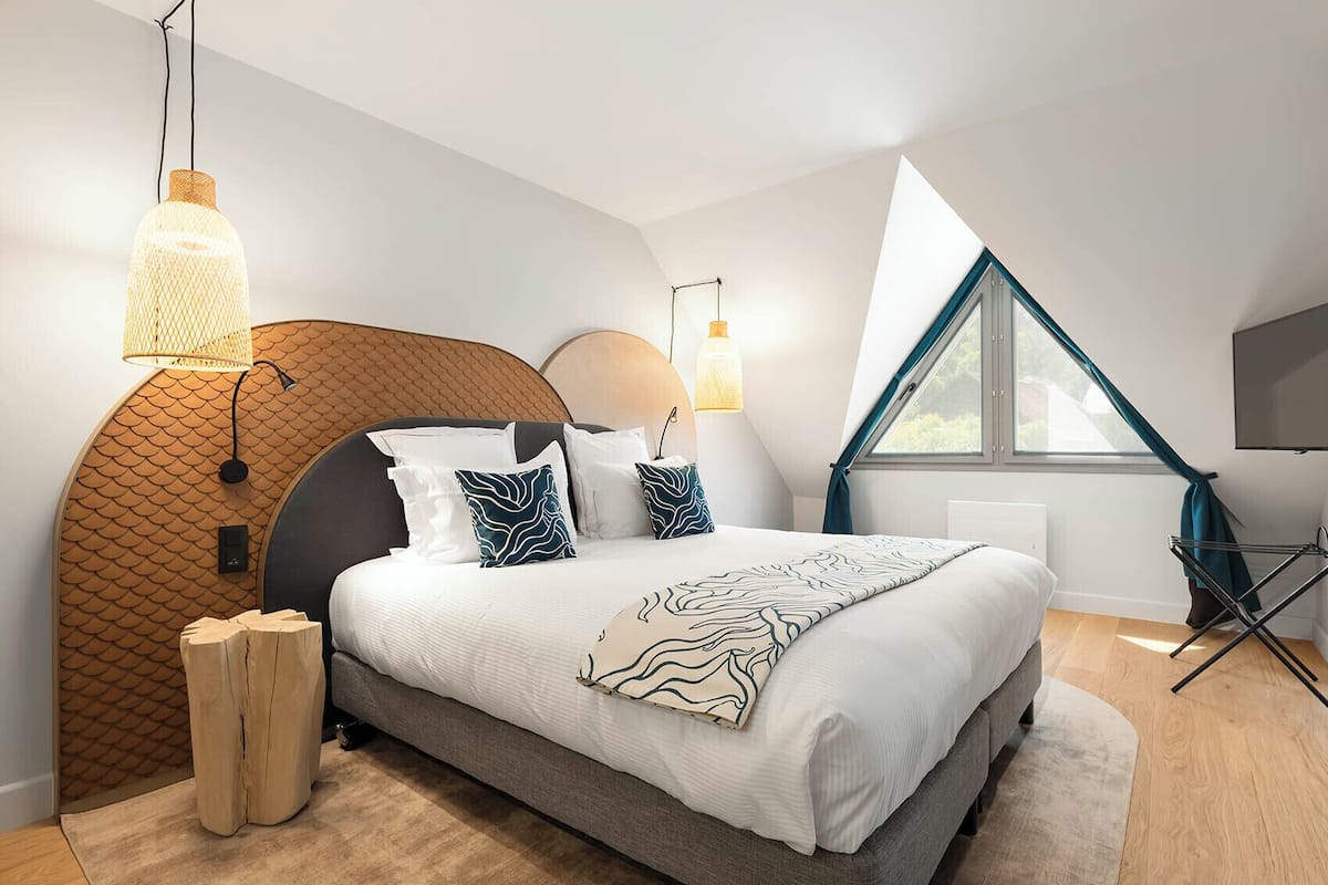 Une chambre à coucher moderne avec une tête de lit incurvée, un grand lit agrémenté d’oreillers à motifs et une fenêtre de toit.