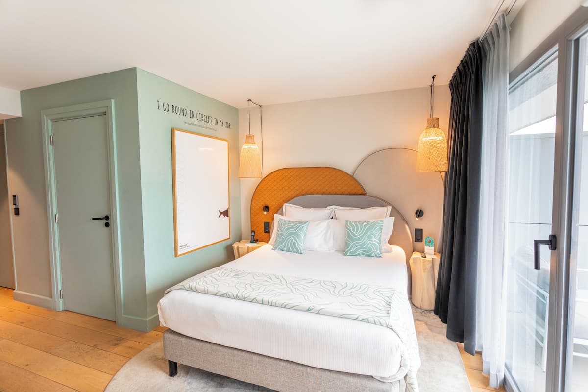 Une chambre à coucher comprenant un lit, une table de chevet, une lampe fixée au mur, une porte et une fenêtre avec des rideaux.