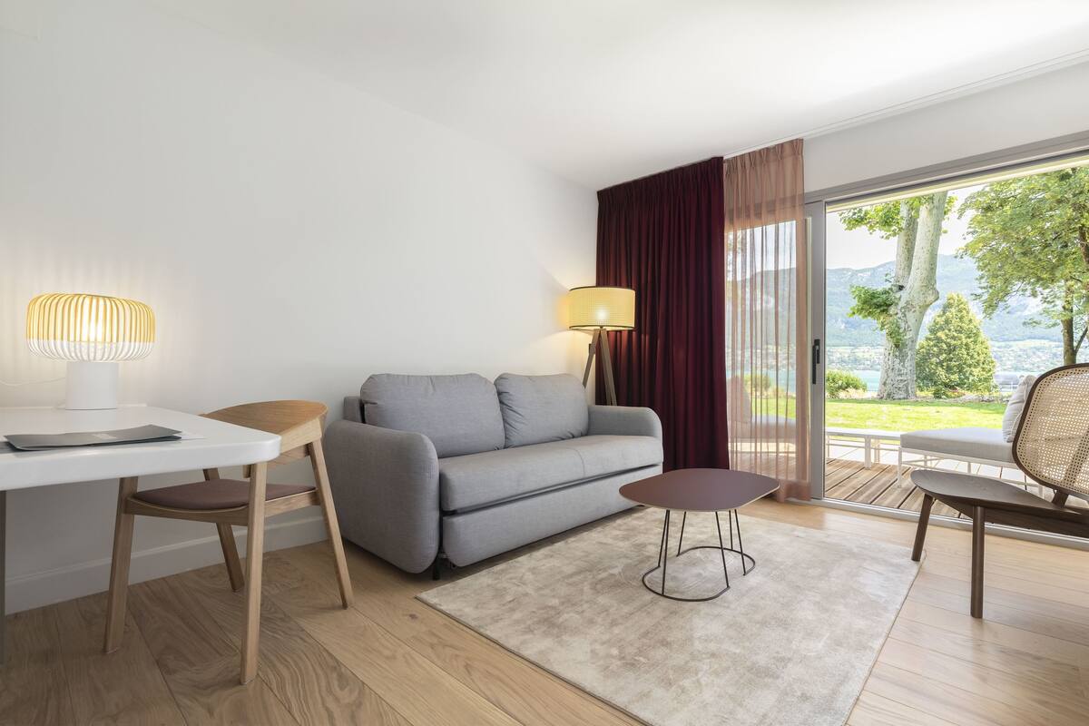 Un salon moderne avec un canapé gris, une table à manger en bois, une petite table ronde et une vue sur un jardin et des montagnes, grâce à de grandes fenêtres.