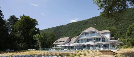 Un hotel moderno con terraza de madera, mobiliario exterior y montañas al fondo.