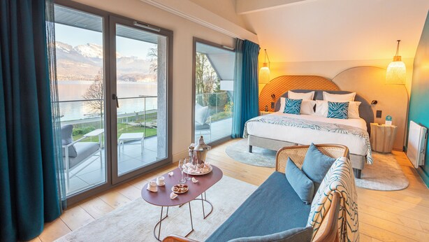 Un dormitorio con cama, sofá, mesa con bandeja de comida y vista a un lago y montañas.