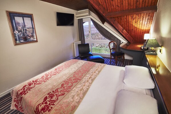 Habitación de hotel con una cama grande, un escritorio con lámpara, una silla, un ventanal con vistas y un cuadro en la pared.