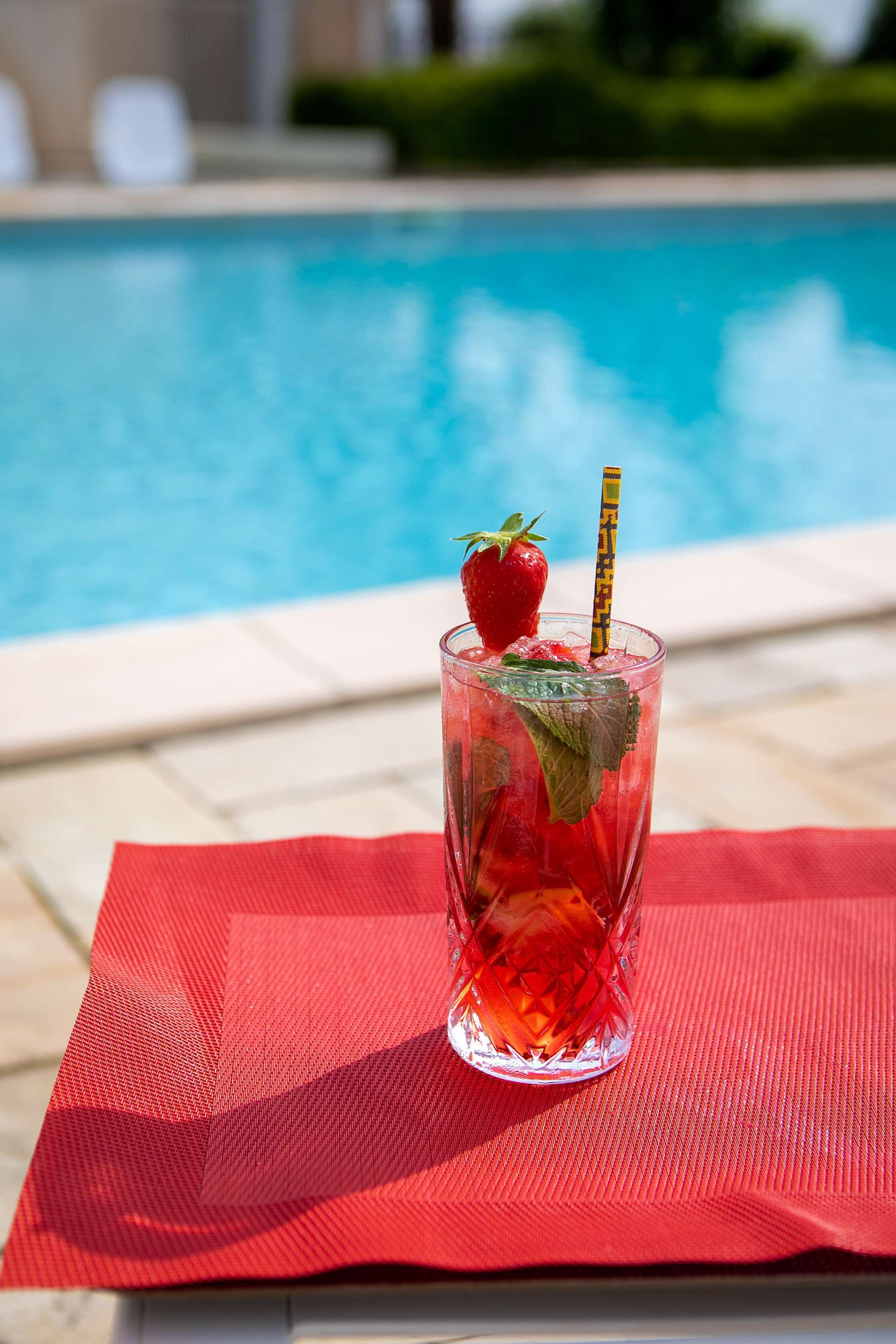 Un vaso de bebida helada con fresa y adornos de menta sobre un tapete rojo junto a una piscina.