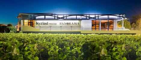 Un bâtiment moderne, sur lequel on peut lire l’enseigne « Kyriad Prestige Panorama », avec un vignoble au premier plan.