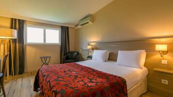 Habitación de hotel con una cama grande, una mesita de noche con lámpara, una silla, una pequeña mesa y una ventana con cortinas.