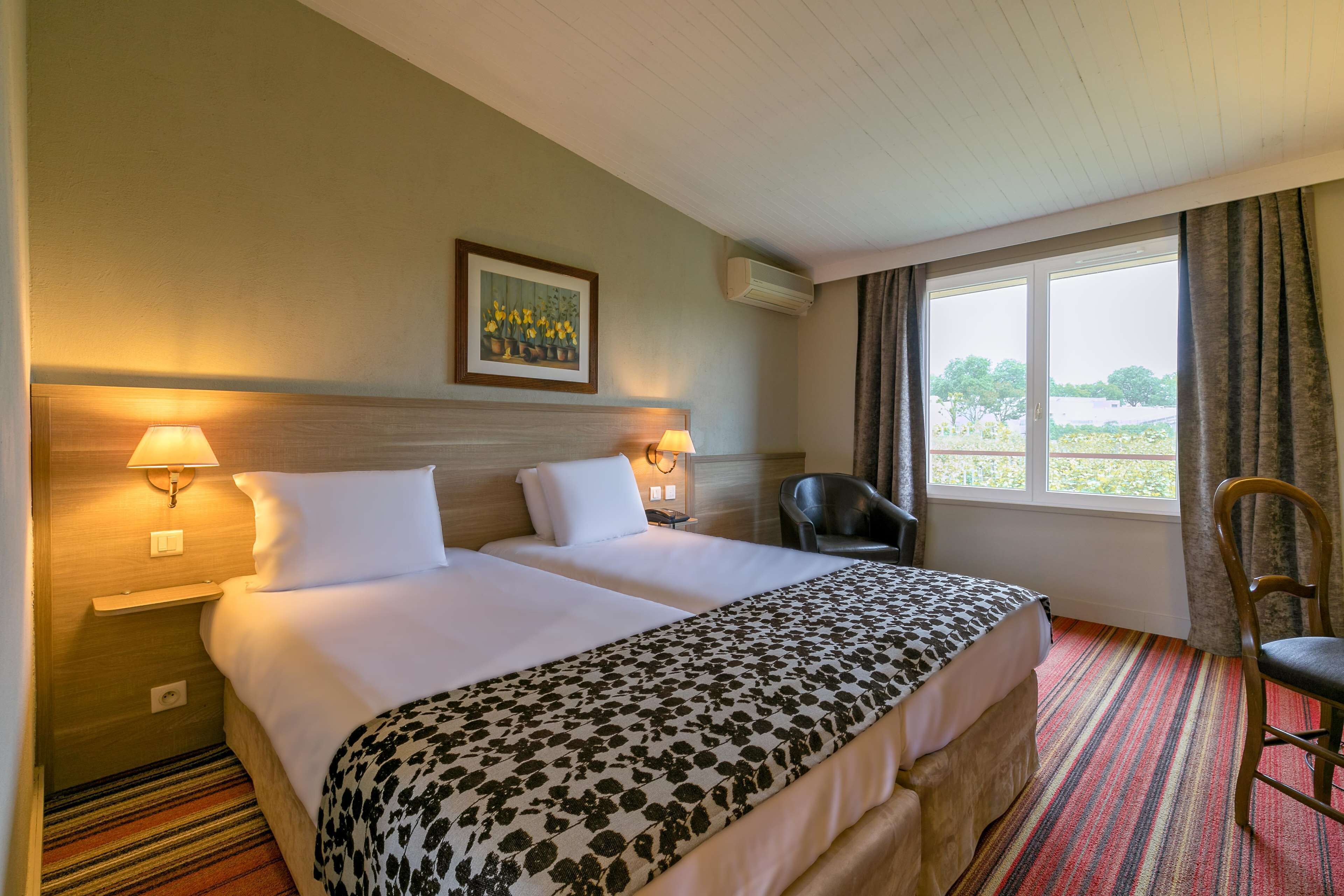 A hotel room with two beds, a chair, a painting, and a window with curtains.