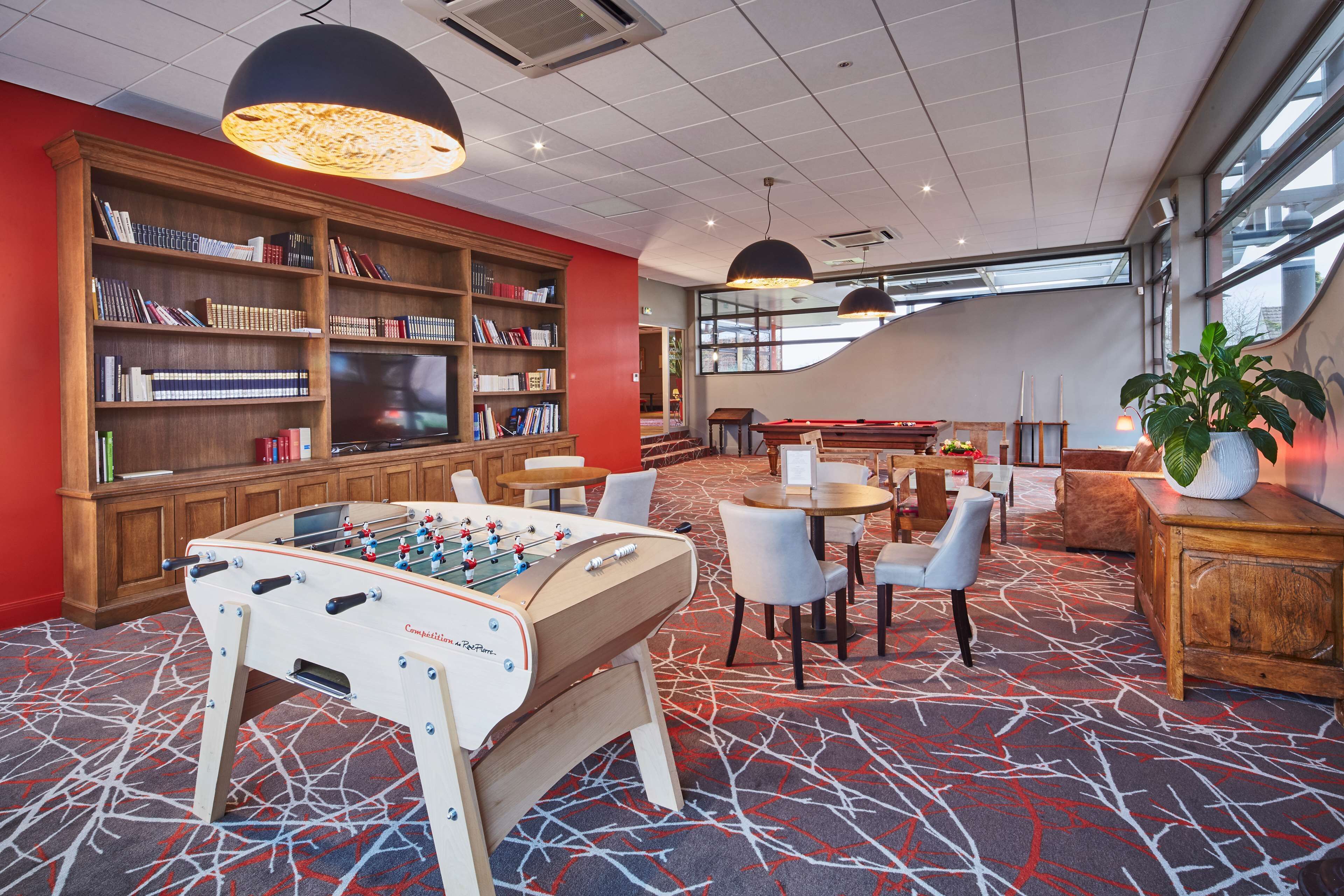 A hotel lobby with a foosball table, a large bookshelf, and a seating area with tables and chairs.