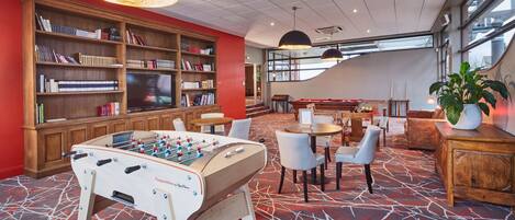 A hotel lobby with a foosball table, a large bookshelf, and a seating area with tables and chairs.