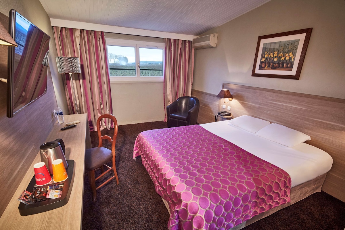 Una habitación de hotel con cama, escritorio con cafetera, silla, lámpara y ventana con cortinas.