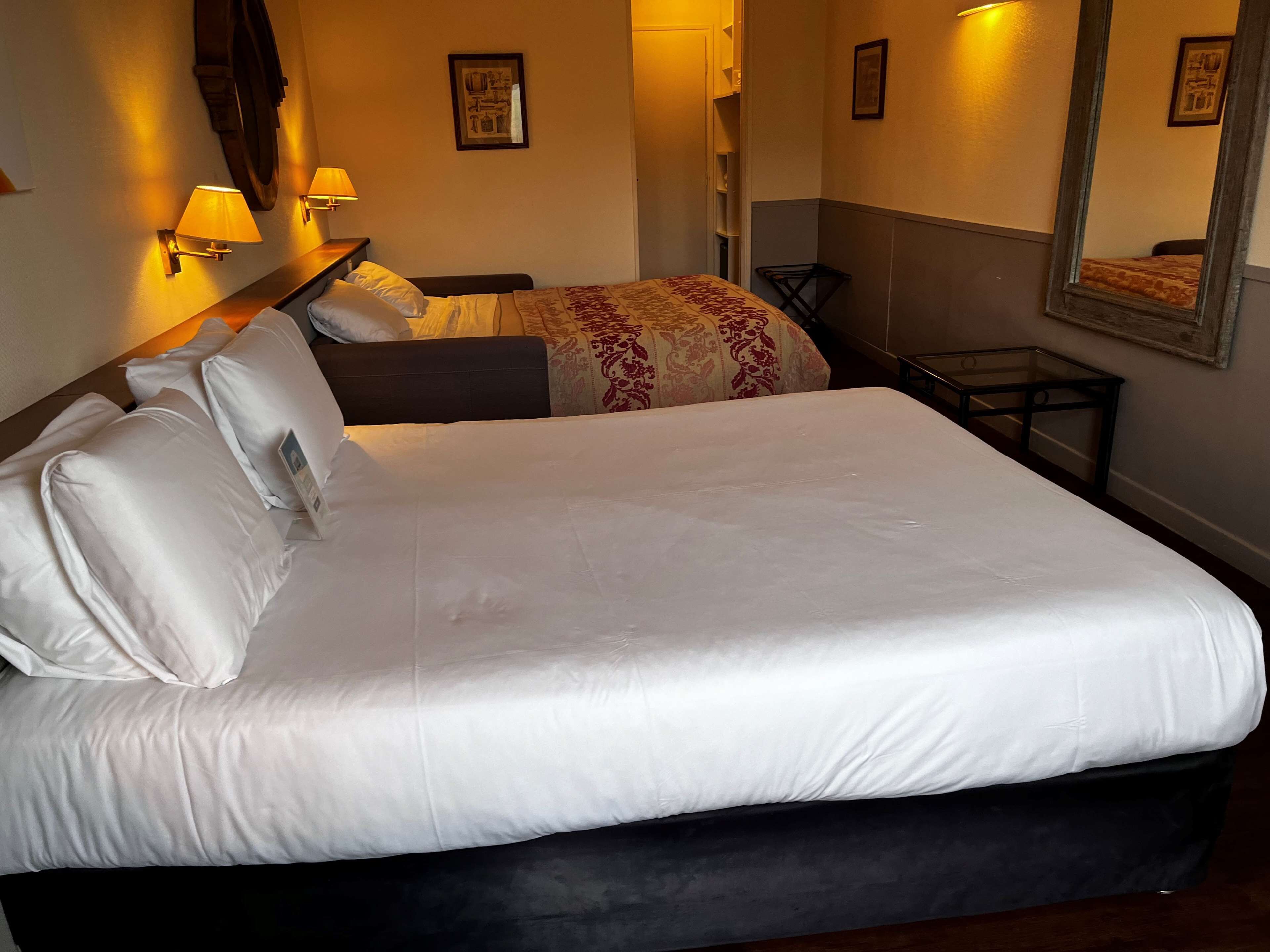 Una cama bien hecha con sábanas y almohadas blancas, una mesita de noche con una lámpara y un espejo pequeño en la pared.