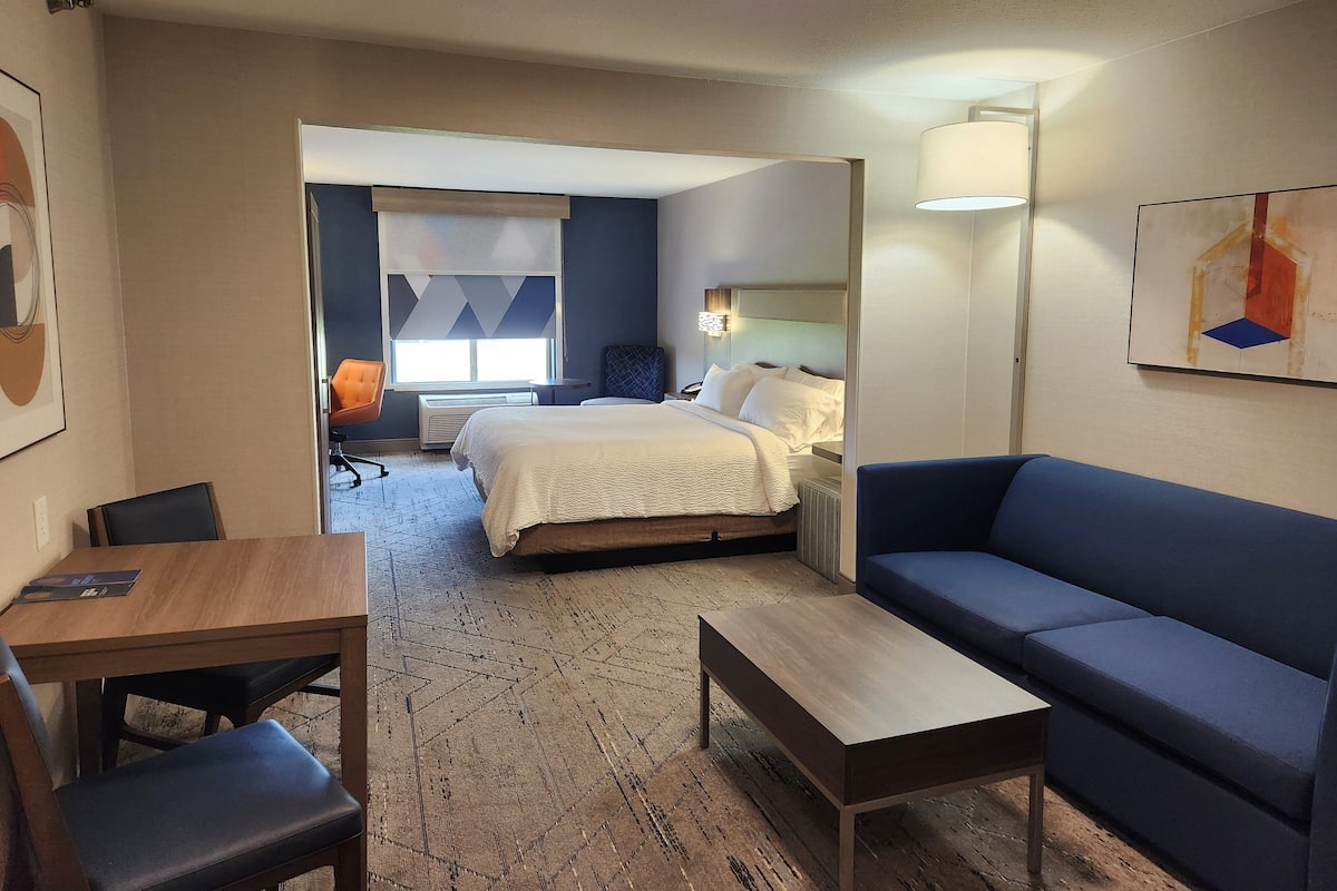 A hotel room with a bed, a blue sofa, a wooden table, a chair, and a desk. There is a window with a view of the outside.