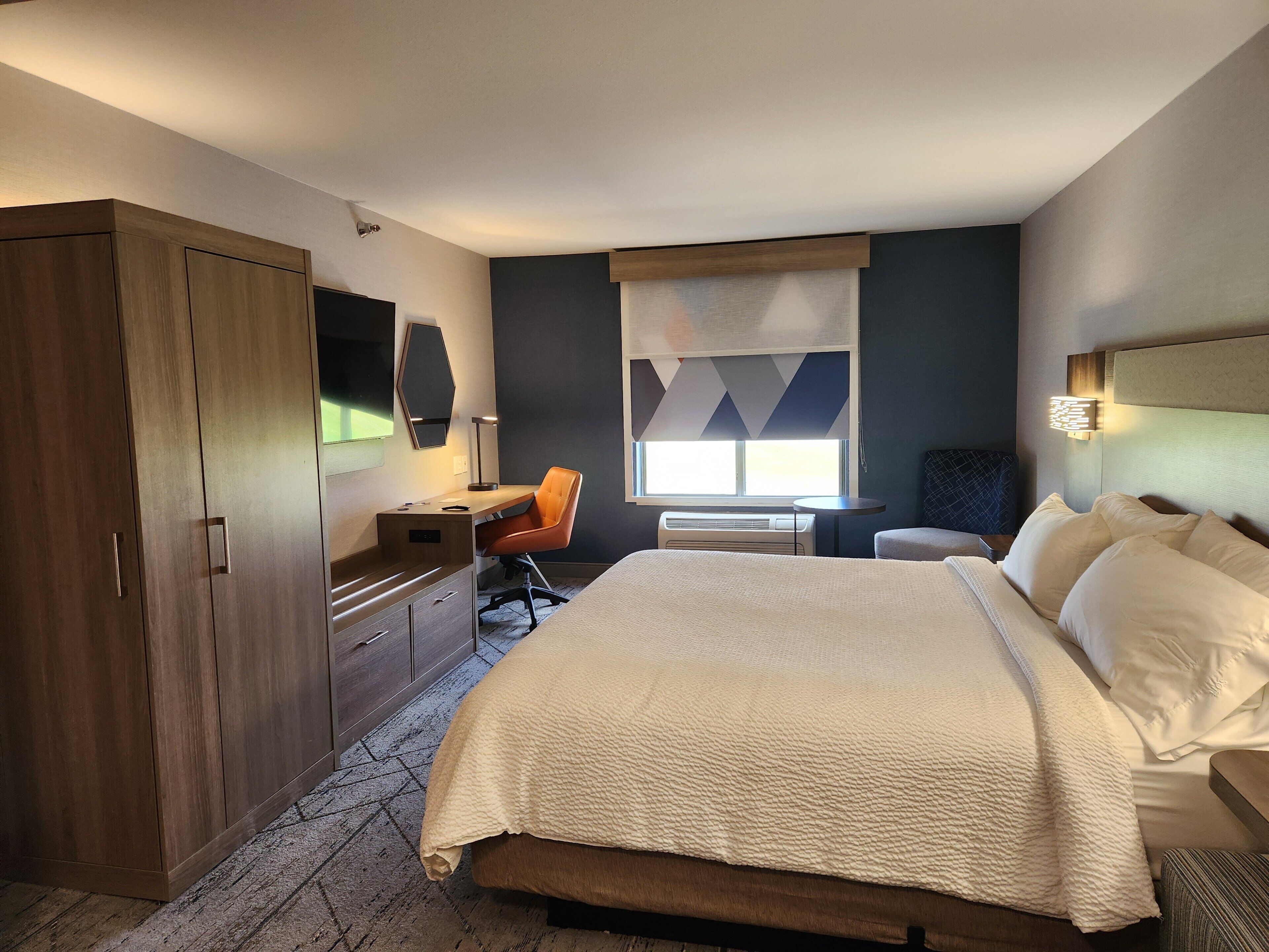 Une chambre d’hôtel avec un grand lit, un bureau avec une chaise, une armoire et une fenêtre avec des stores.