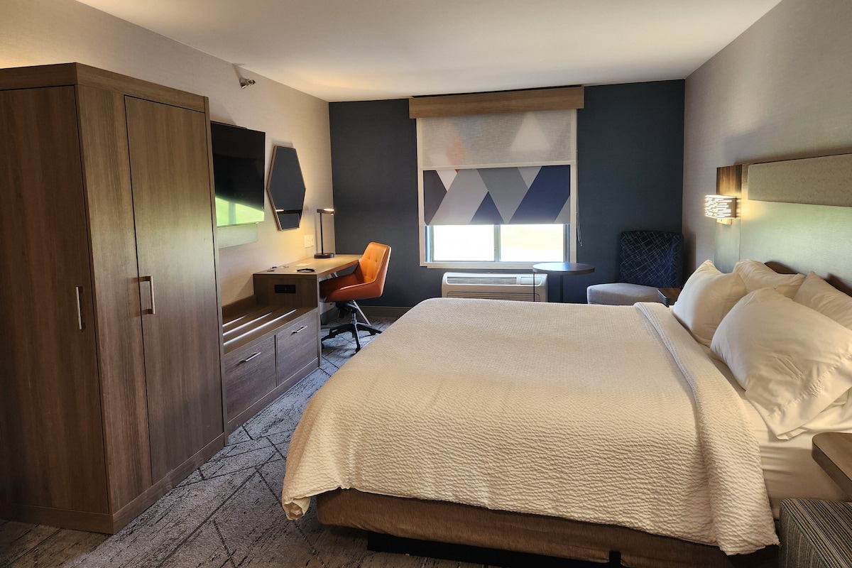 A hotel room with a bed, a desk, a chair, a wardrobe, and a window with blinds.