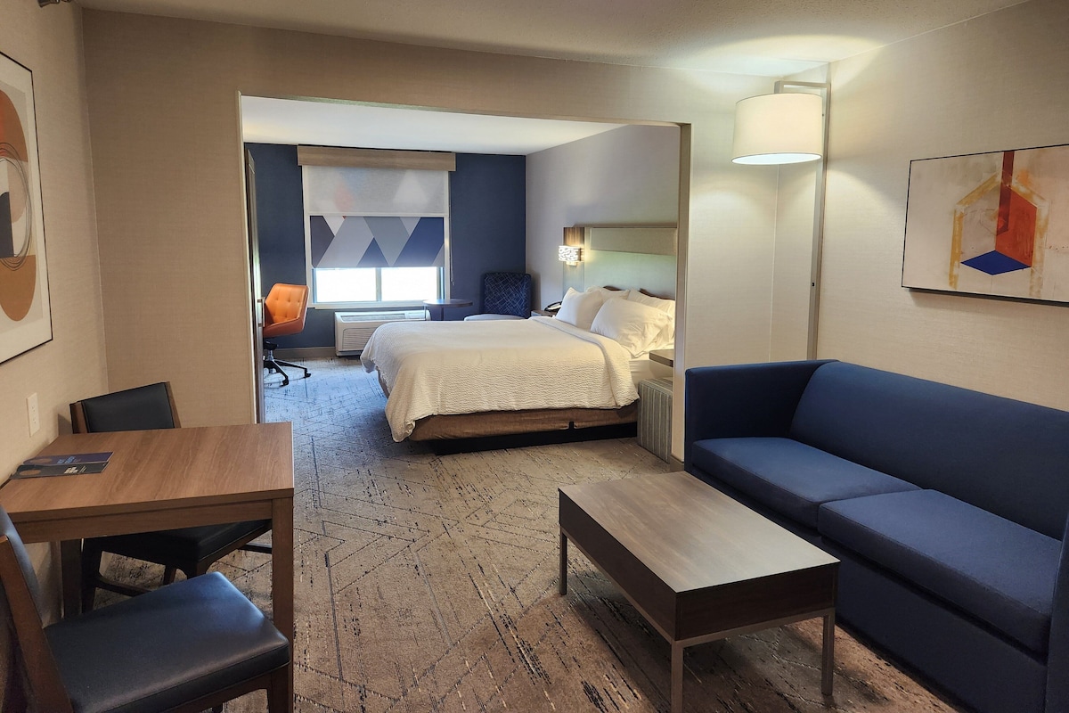 A hotel room with a bed, a blue sofa, a wooden table, a chair, and a desk. There is a window with a view of the outside.