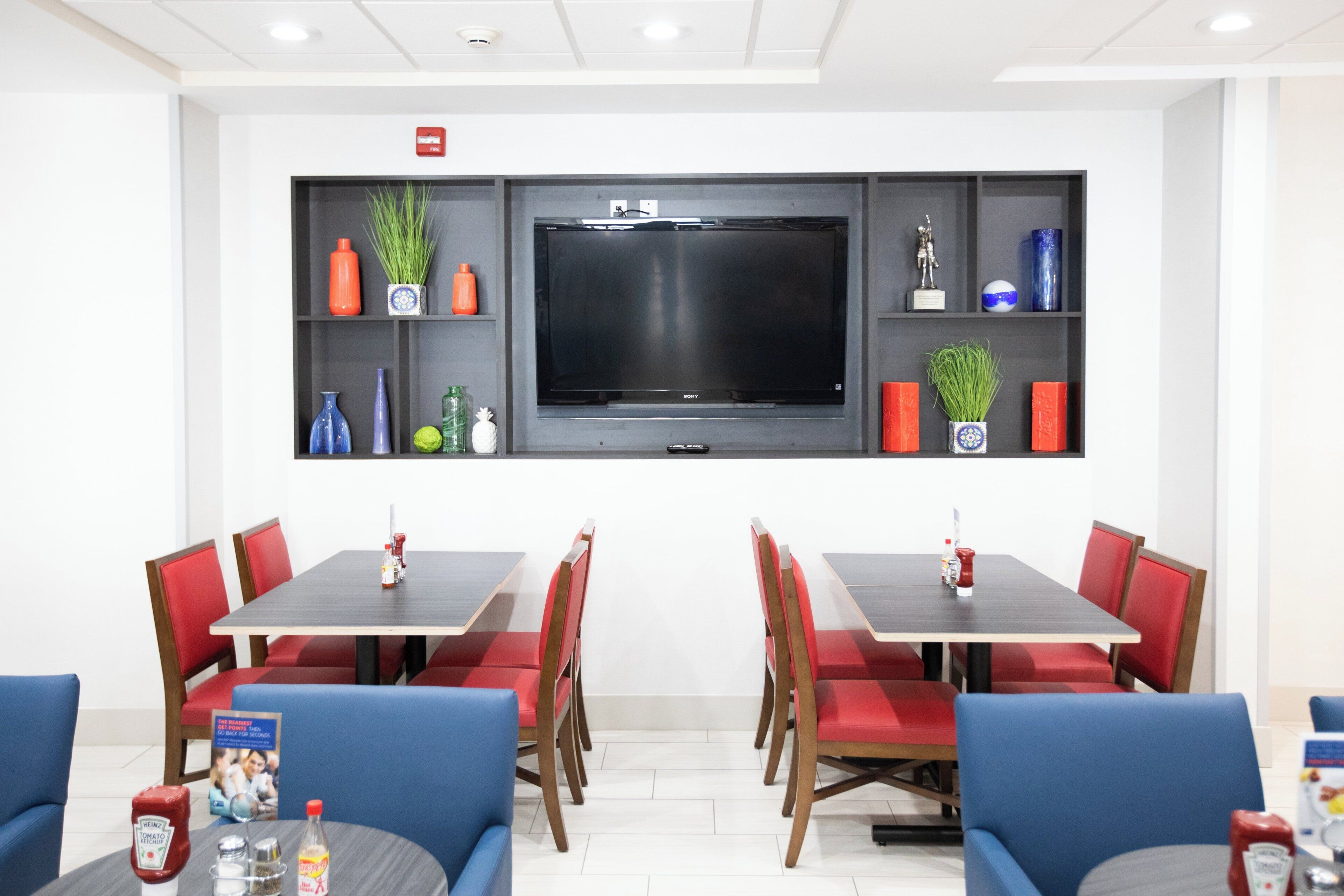 A conference room with a large flat-screen TV, red and blue chairs, and a white table.