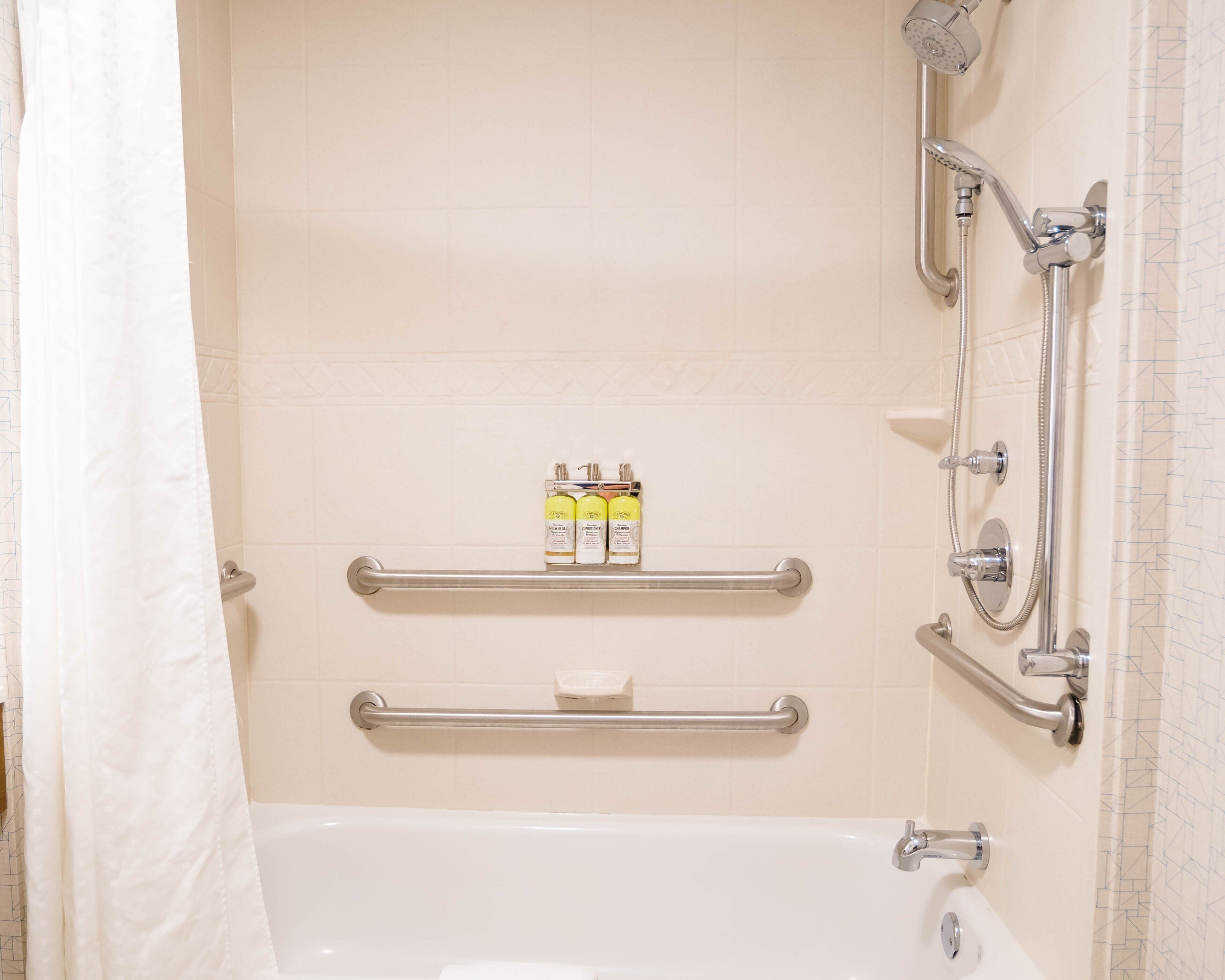 Une salle de bain équipée d’une baignoire, d’une douche avec un pommeau de douche amovible et de deux barres d’appui.